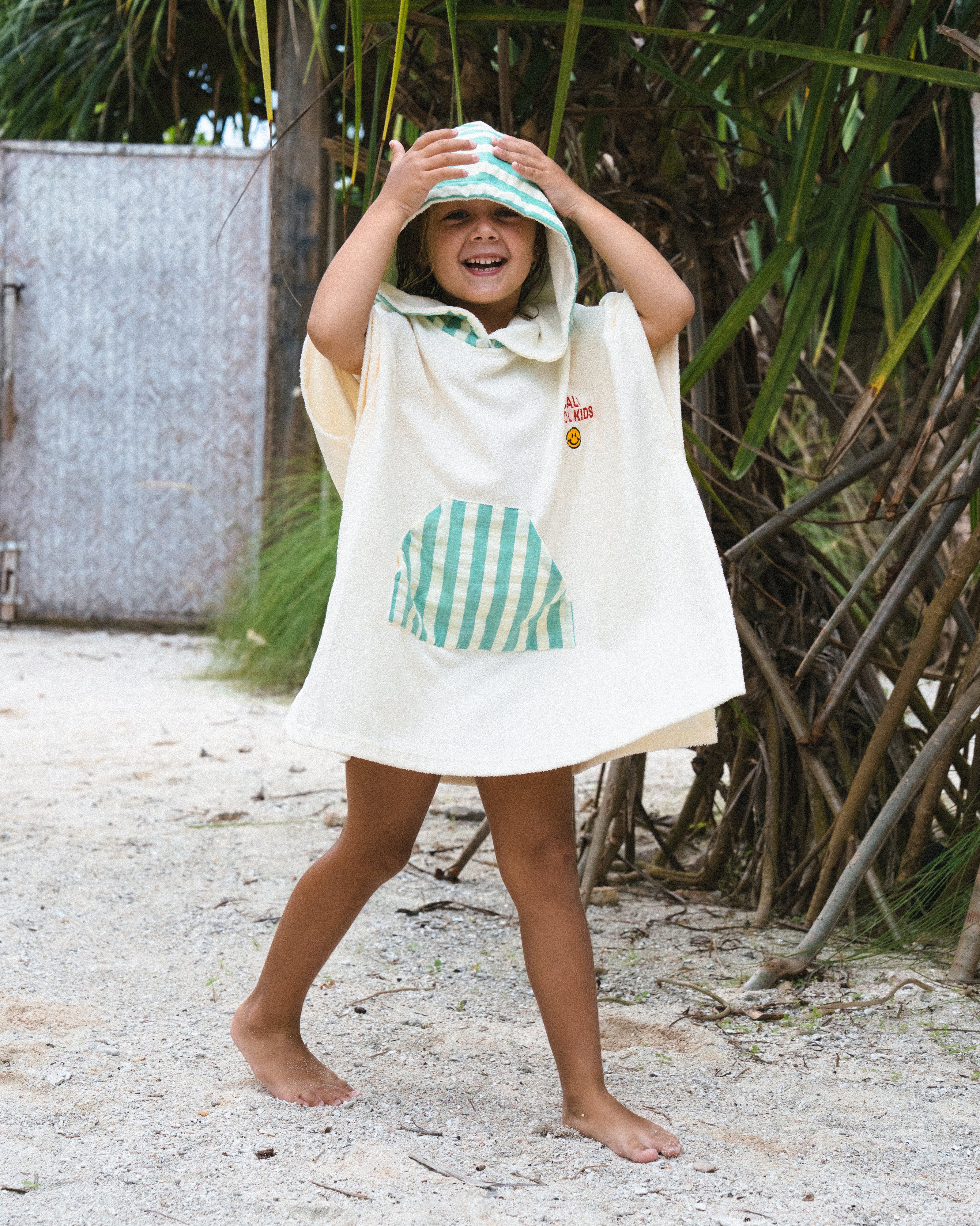 A child wearing a Club Pantai white poncho and a hood, standing on a sandy beach.
