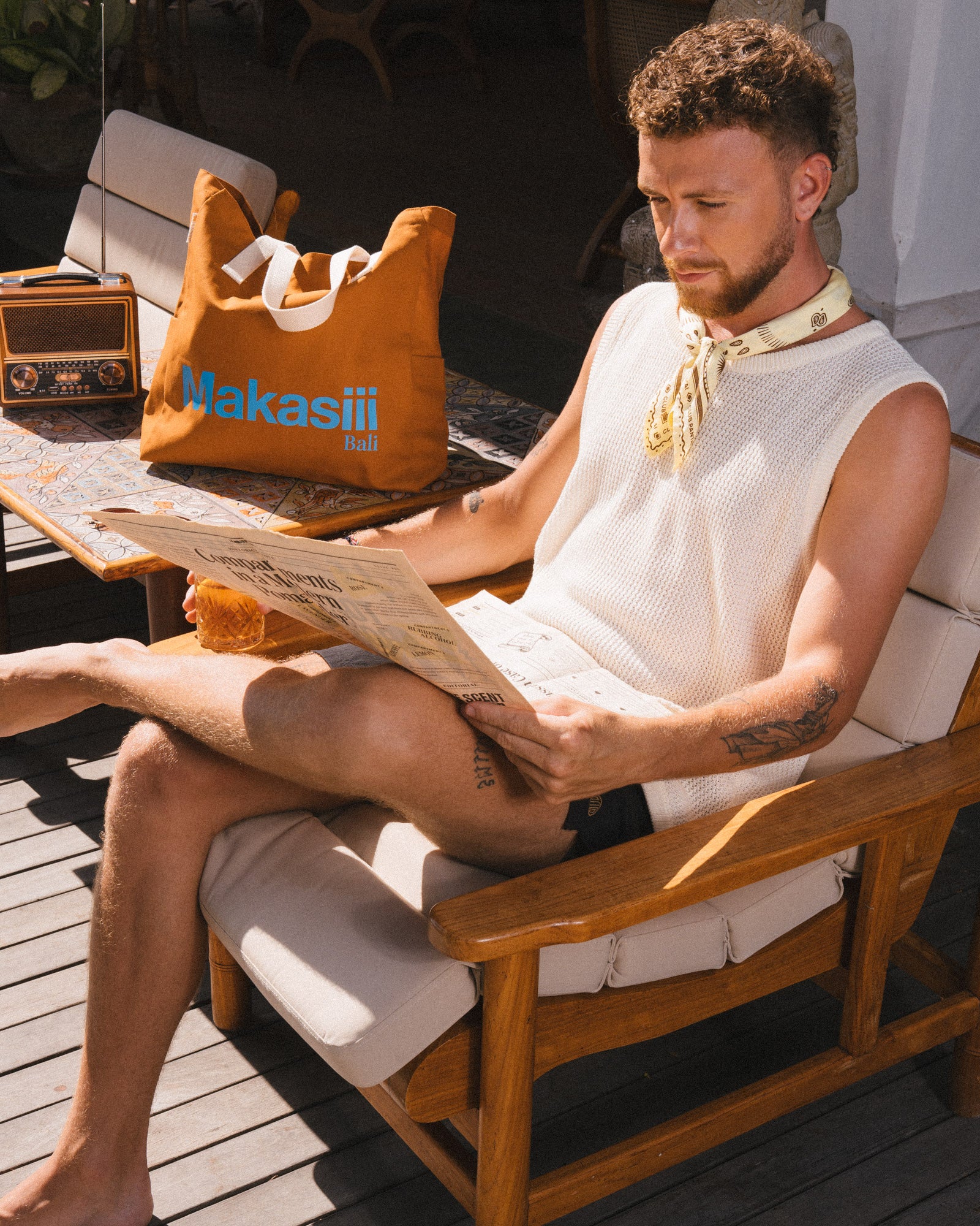 Man reading a newspaper on a wooden chair with a Makasii bag and a yellow bandana from Club Pantai