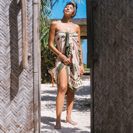 A woman in a sarong padang padang from Club Pantai, standing in a tropical setting with palm trees.